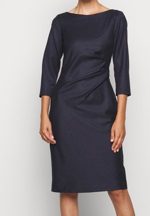Kobieta ubrana w dopasowaną, granatową sukienkę do kolan z rękawami do połowy przedramienia i dekoltem typu łódka, stojąca na jednolitym tle.