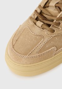 Suedesk sneaker i lys tan med rund tå, teksturert overdel og flate lisser. Gummisålen har et teksturert mønster for grep.