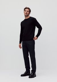 Zwarte ronde hals trui, zwarte slim-fit broek en zwarte leren schoenen. Model staat op een effen achtergrond, handen op de heupen.