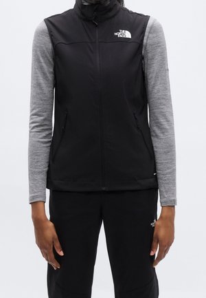 Person trägt eine schwarze North Face Weste über einem grauen Langarmshirt und schwarzen Hosen, die Hände entspannt an den Seiten.