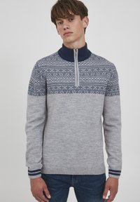 Lys grå strikkegenser med marineblå detaljer, med en glidelås i kragen og geometriske mønstre på brystet. Ribbestrikkede mansjetter og skjorte.