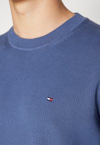 Gros plan sur une personne portant un pull en maille bleu avec un petit logo rectangulaire rouge, blanc et bleu sur la poitrine.