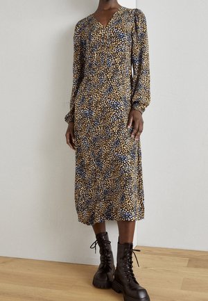 Femme portant une robe midi à manches longues et col en V avec un motif bleu, marron et blanc, associée à des bottines noires à lacets sur un sol en bois.