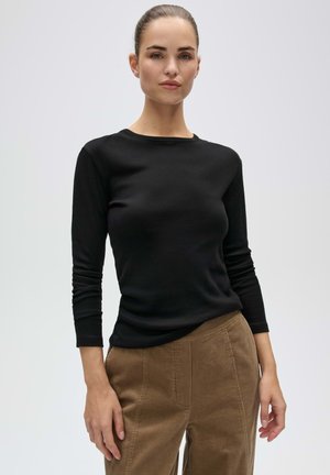 Femme aux cheveux attachés portant un haut noir à manches longues et un pantalon en velours côtelé marron, debout devant un fond clair uni.