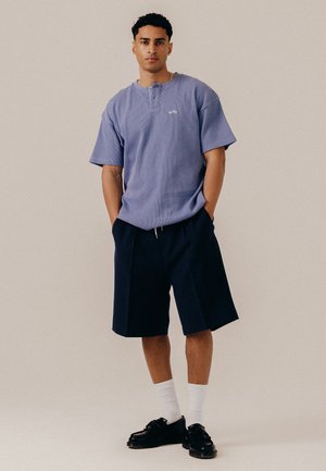 Ung mand, der står med hænderne i lommerne iført en lilla kortærmet skjorte, marineblå shorts, hvide sokker og sorte sko på en ensfarvet baggrund.