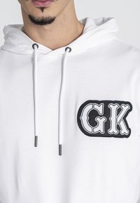 Sudadera de algodón blanca con un logotipo bordado en negro "GK" en el lado izquierdo del pecho, con cordones y detalles metálicos. Textura suave.