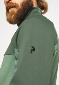 Groene sportjack met een gestructureerd accent op de schouder en een zwart logo op de mouw. Glad materiaal met een getailleerd ontwerp.