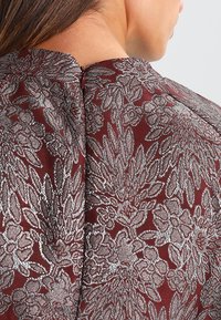 Burgundská tkanina s složitou kovovou stříbrnou květinovou výšivkou, s vysokým výstřihem a mírným rozparkem vzadu. Hladký, texturovaný povrch.