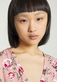 Vestido floral rosa com um design de decote em V. O tecido apresenta rosas vermelhas e verdes sobre um fundo suave, com cabelo curto preto emoldurando o rosto.