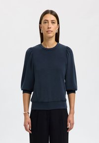 Pullover blu scuro con maniche a palloncino, scollo tondo e orlo a costine. Realizzato in tessuto liscio con una texture morbida. Indossato con pantaloni neri.