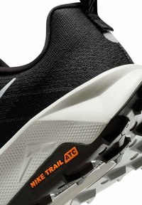 Svart texturerad textilöverdel med en grå och vit gummisula. Har "Nike Trail ATC"-logotyp i orange och distinkt greppmönster.