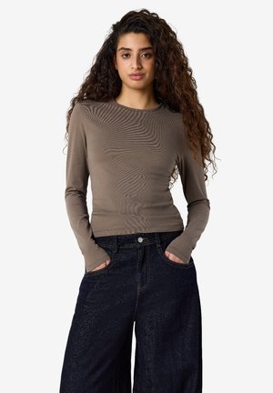 Ung kvinde med langt krøllet hår iført en tætsiddende brun langærmet skjorte og mørke vidde jeans med hænderne i lommerne.