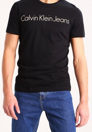 Schwarzes kurzärmeliges T-Shirt mit weißem "Calvin Klein Jeans"-Schriftzug über der Brust, getragen mit blauen Denim-Jeans.