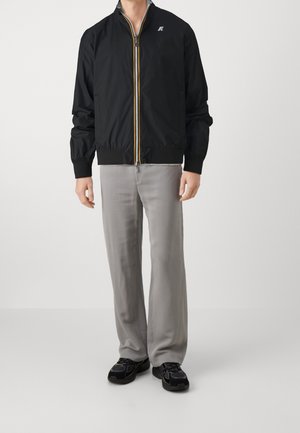 Mand iført sort lynlåstrøje, lysegrå løse bukser og sorte sneakers, stående mod en ensartet hvid baggrund.