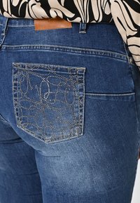 Jean en denim bleu foncé avec une poche texturée ornée d'un motif de perles argentées décoratives. Comprend une étiquette en cuir marron au niveau de la taille.