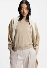 Cremefarvet sjal lagd over en beige langærmet bluse, parret med lyse beige sålede bukser. Bløde teksturer og neutral farvepalet.