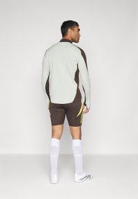 Haut sportif à manches longues en gris clair et marron, associé à un short marron avec des accents jaunes, porté avec des chaussettes jusqu'aux genoux blanches.
