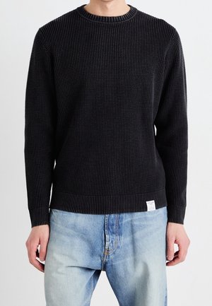 Man wearing a black textured knit sweater and light blue faded jeans, standing against a plain white background.