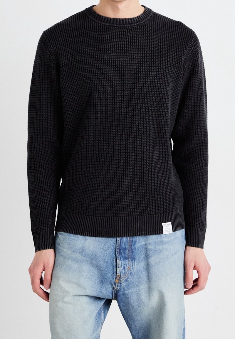 Homme portant un pull en maille noire texturée et un jean bleu clair délavé, debout devant un fond blanc uni.