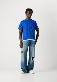 Joven vestido con una camiseta azul brillante, jeans anchos claros rasgados y zapatillas blancas, de pie frente a un fondo blanco liso.