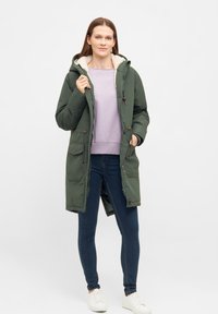 Grön vadderad parkas med huva, med en mjuk krämfärgad foder, tryckknapp- och dragkedjestängning samt fyra framfickor. Bärs över en lila topp.
