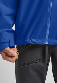 Veste imperméable bleue avec fermeture éclair et ourlet ajustable, associée à un pantalon noir avec une poche zippée et des détails de couture grise.