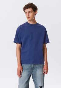 Kortærmet, rundhalset t-shirt i solidt mørkeblåt stof, lavet af bomuld, med en afslappet pasform og blød tekstur. Bæres sammen med lyseblå slidte jeans.