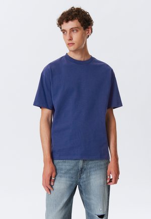 T-shirt à manches courtes et col rond en tissu bleu foncé uni, en coton, avec une coupe décontractée et une texture douce. Porté avec un jean distressed bleu clair.