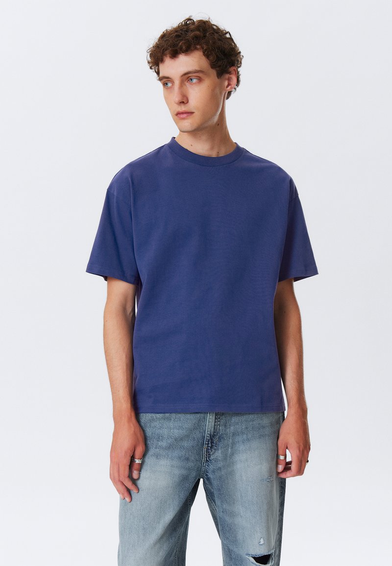 Kortærmet, rundhalset t-shirt i solidt mørkeblåt stof, lavet af bomuld, med en afslappet pasform og blød tekstur. Bæres sammen med lyseblå slidte jeans.