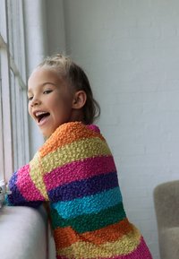 Multicolored knit sweater with horizontal stripes in orange, yellow, pink, purple, turquoise, and green; soft texture; worn by a child.