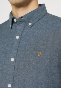 Modrá košile chambray s límečkem na knoflíky, bílými knoflíky a malým žlutě vyšitým logem na hrudi. Hladká textura.