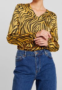 Persona che indossa una blusa a righe gialle e nere in stile tigre, infilata in jeans di denim blu a vita alta, con le mani unite in vita.