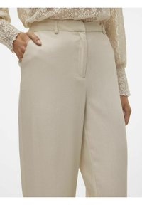 Pantalons beiges à taille haute, texture lisse, devant plissé et poches latérales. Associés à un blouse en dentelle à manches longues.