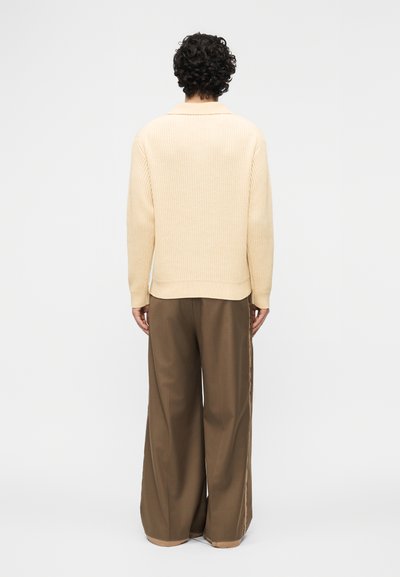 Personne aux cheveux bouclés, vue de dos, portant un pull en tricot crème et un pantalon marron à larges jambes, sur fond blanc uni.