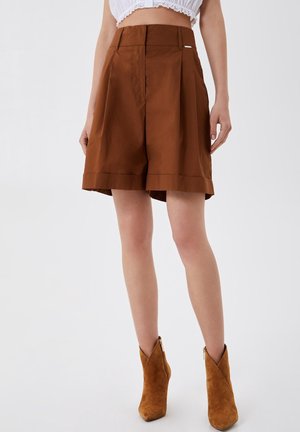 Femme portant un short marron taille haute et des bottines en daim beige, debout devant un fond blanc uni.