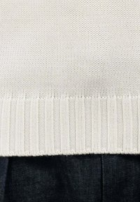 Maglione lavorato a maglia in bianco sporco, con orlo a coste e motivo testurizzato; indossato sopra un tessuto di denim scuro con pieghe.
