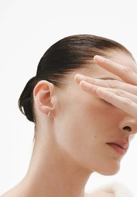 Pendiente de plata con un diseño angular y superficie texturizada. El cabello del modelo está peinado hacia atrás y su mano oculta parcialmente su rostro.