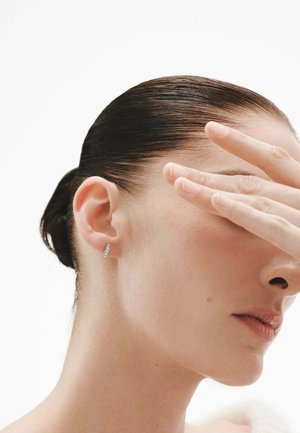 Pendiente de plata con un diseño angular y superficie texturizada. El cabello del modelo está peinado hacia atrás y su mano oculta parcialmente su rostro.