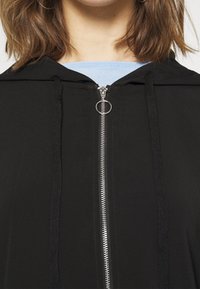 Svart zip-up hoodie med silverfärgad dragkedja, huva och dragsnoddar. Tyget ser glatt och lätt ut, med en lager-ljusblå tröja under.