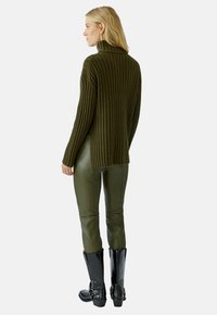 Olivefarbener Rippstrickpullover mit hohem Kragen, kombiniert mit olivgrünen Lederhosen und schwarzen knielangen Stiefeln mit Schnallen.