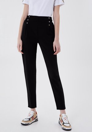 Osoba ubrana w czarne spodnie z wysokim stanem z ozdobnymi guzikami oraz wielokolorowe buty sportowe w białym, beżowym i czarnym kolorze, stojąca na tle jednolitego tła.