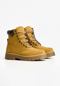 Gelbe Wildleder-Wanderschuhe mit braunen Akzenten, Metallösen und strukturierten Schnürsenkeln. Dicke Gummisohlen mit einem geriffelten Muster für besseren Halt.