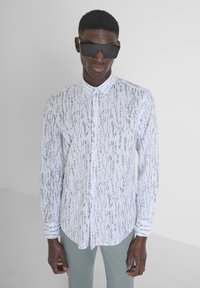 Camicia bianca con un motivo tessurato in grigio, dotata di colletto e chiusura con bottoni. Il modello indossa occhiali da sole neri angolari.