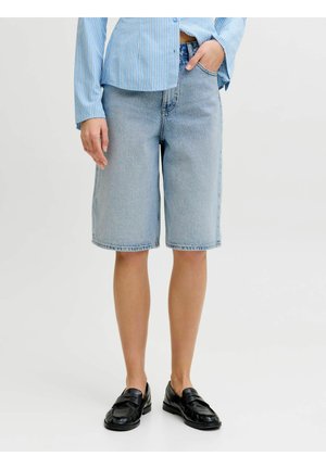 Person iført lyseblå denim knælange shorts, en blåstribet skjorte med knapper og sorte loafers, stående med den ene hånd i lommen.