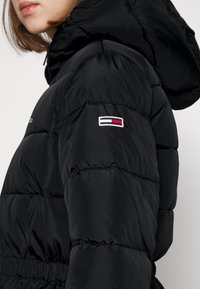 Černá péřová bunda s kapucí, s prošívanými sekcemi a malým vyšitým logem na rukávu v červené, bílé a modré barvě.