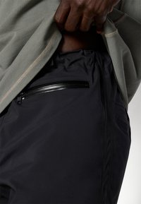 Mustat housut, joissa on rakennekangasta, tyylikäs vetoketjullinen tasku ja joustava vyötärö. Yhdistetty sagevihreään puseroon, jossa on kontrastiväriset tikkaukset.