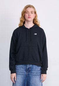 Svart zip-up hettegenser i mykt stoff, med en frontlomme, snor i hetten og en hvit logo på venstre bryst. Kombinert med lyseblå jeans.