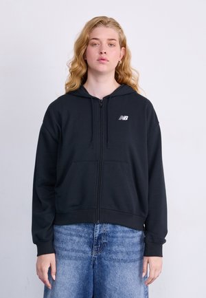 Zwarte rits hoodie van zachte stof, met een voorkant zak, capuchon met trekkoord en een wit logo op de linkerborst. Gecombineerd met lichtblauwe jeans.