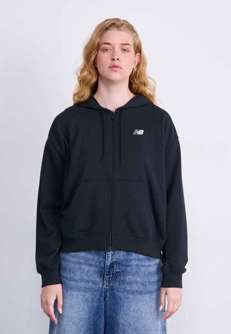 Svart zip-up hettegenser i mykt stoff, med en frontlomme, snor i hetten og en hvit logo på venstre bryst. Kombinert med lyseblå jeans.