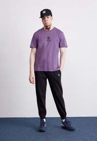 Violeta kokvilnas T-krekls ar melnu Timberland logo, savienots ar melnajām sporta biksēm un tumšpelēkiem apaviem. Melna cepure noslēdz apģērbu.